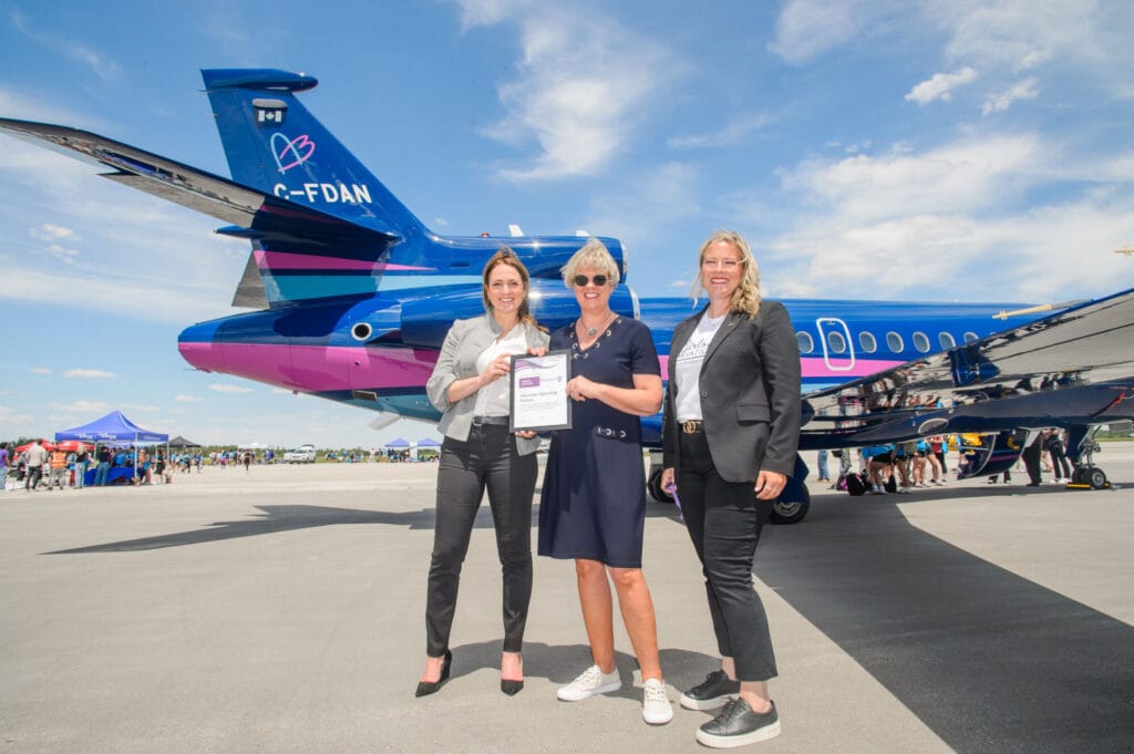 Laura Briscoe, Florentine Strzelczyk, and Jackie Book stand on the tarmac at London International Airport holding a partnership certificate in front of a blue and pink Aubrey Dan’s aircraft during Girls in Aviation Day.