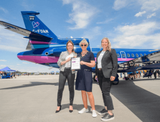 Laura Briscoe, Florentine Strzelczyk, and Jackie Book stand on the tarmac at London International Airport holding a partnership certificate in front of a blue and pink Aubrey Dan’s aircraft during Girls in Aviation Day.