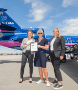 Laura Briscoe, Florentine Strzelczyk, and Jackie Book stand on the tarmac at London International Airport holding a partnership certificate in front of a blue and pink Aubrey Dan’s aircraft during Girls in Aviation Day.
